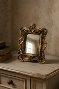 Miroir d'appoint, baroque doré 13×16 cm, posé sur un bureau en bois patiné parmi des livres anciens.